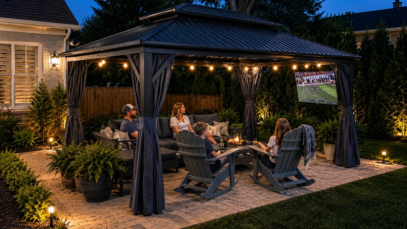 A large dark gray outdoor patio hardtop gazebo with string lights and four people sitting underneath on chairs and a sofa, watching a soccer game on a screen mounted on the side. The scene is set in a landscaped backyard at dusk.