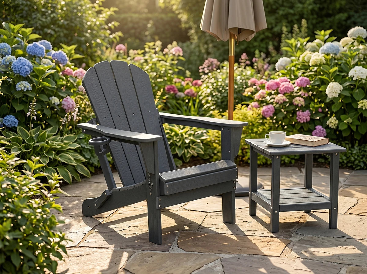 A dark gray outdoor Adirondack chair sits on a stone patio beside a matching side table, with a closed patio umbrella, flowers, and greenery in the background.#color_Slate Gray