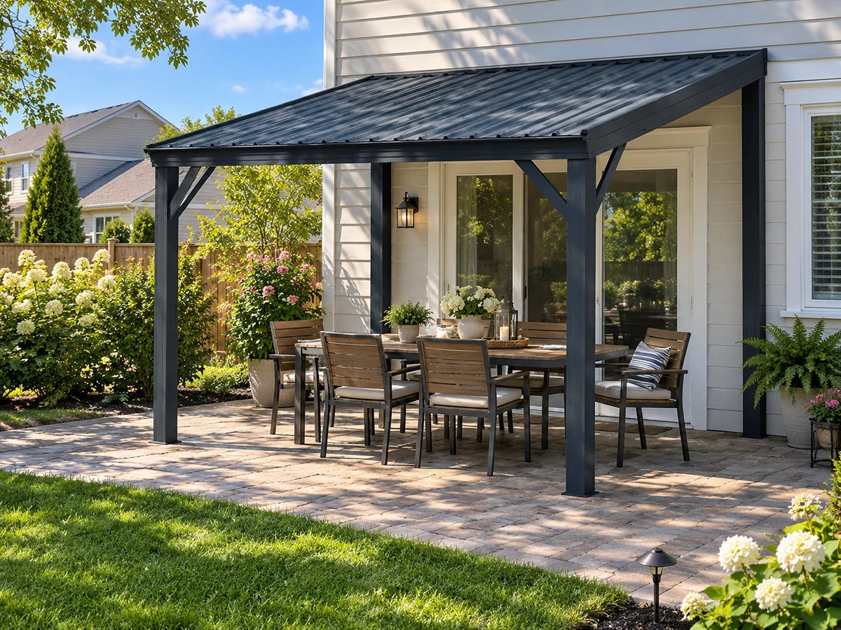 A black metal patio cover with a matching outdoor dining table and chairs, set on a paved backyard patio in front of a white house with green landscaping.