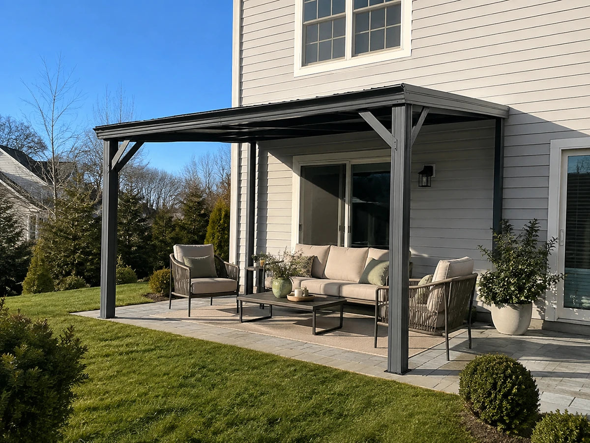 A dark gray metal patio cover canopy installed over a covered porch with a beige outdoor sofa, two armchairs, and a coffee table. The porch is attached to a house with light gray siding, and the surrounding area features a green lawn and landscaping under a clear blue sky.