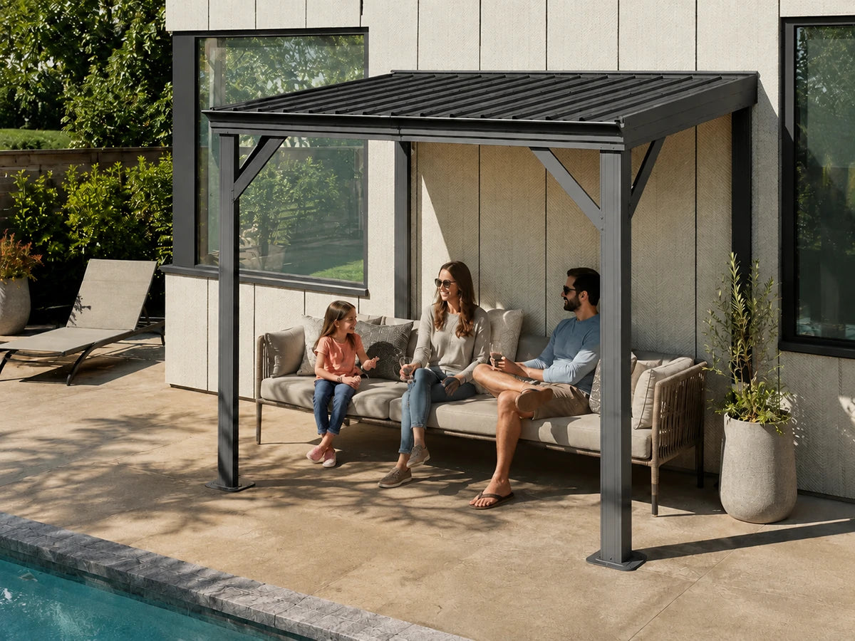 A modern outdoor patio gazebo with a dark gray metal frame and slatted roof, sheltering a beige cushioned sofa where a family of three is sitting. The cabana is situated next to a swimming pool on a stone patio, with a contemporary house in the background.