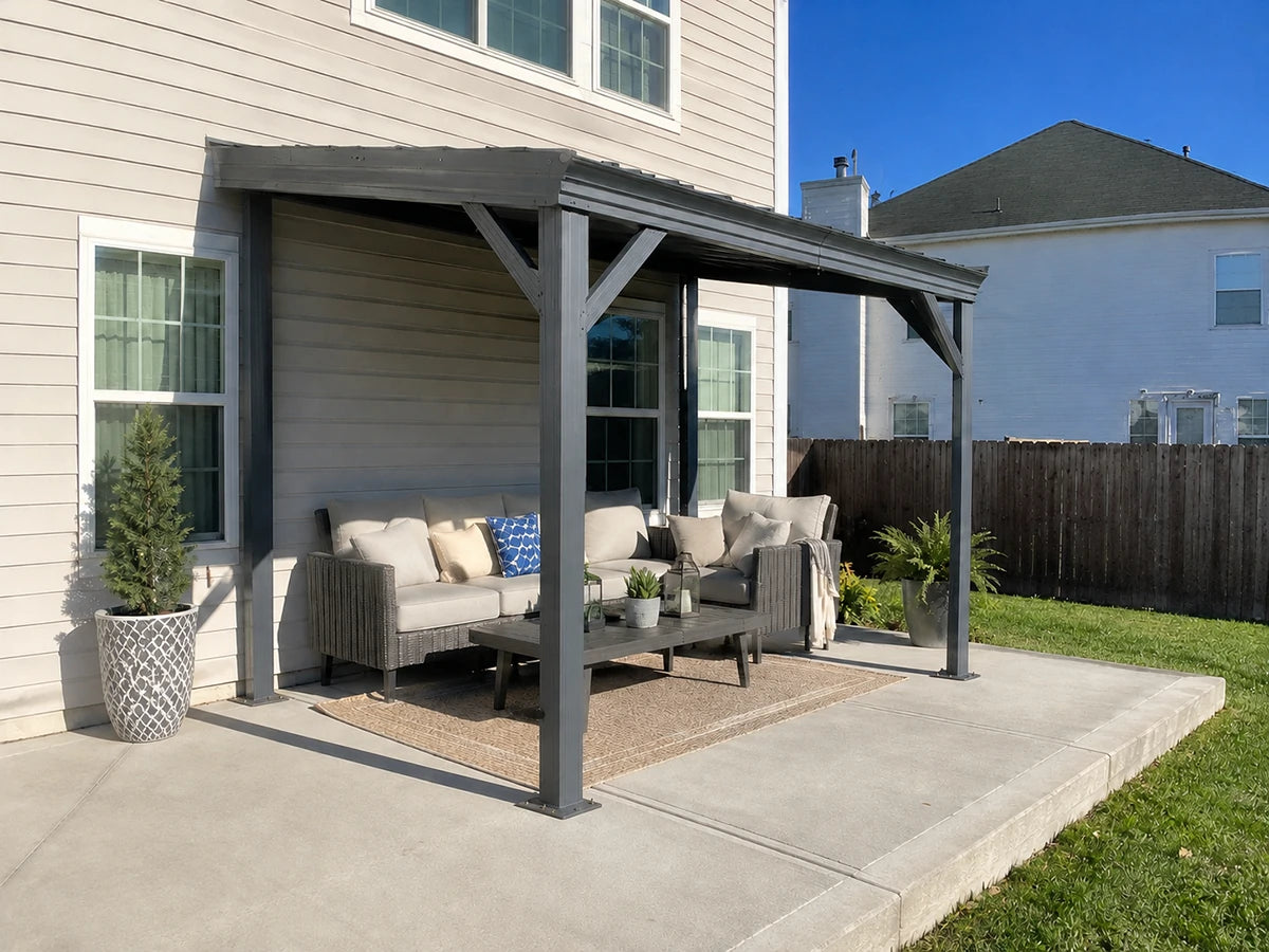 A dark gray outdoor patio cover with a built-in seating area, shown installed on a concrete patio in a backyard setting next to a beige house.